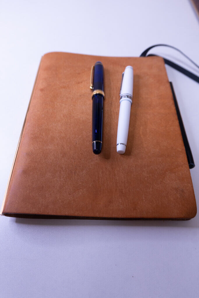 キャメル色のプロッター A5サイズの革表紙の上に『プラチナ万年筆 ＃3776 センチュリー(青)』と『セーラー万年筆 プロギアスリム(白)』の万年筆を並べた写真